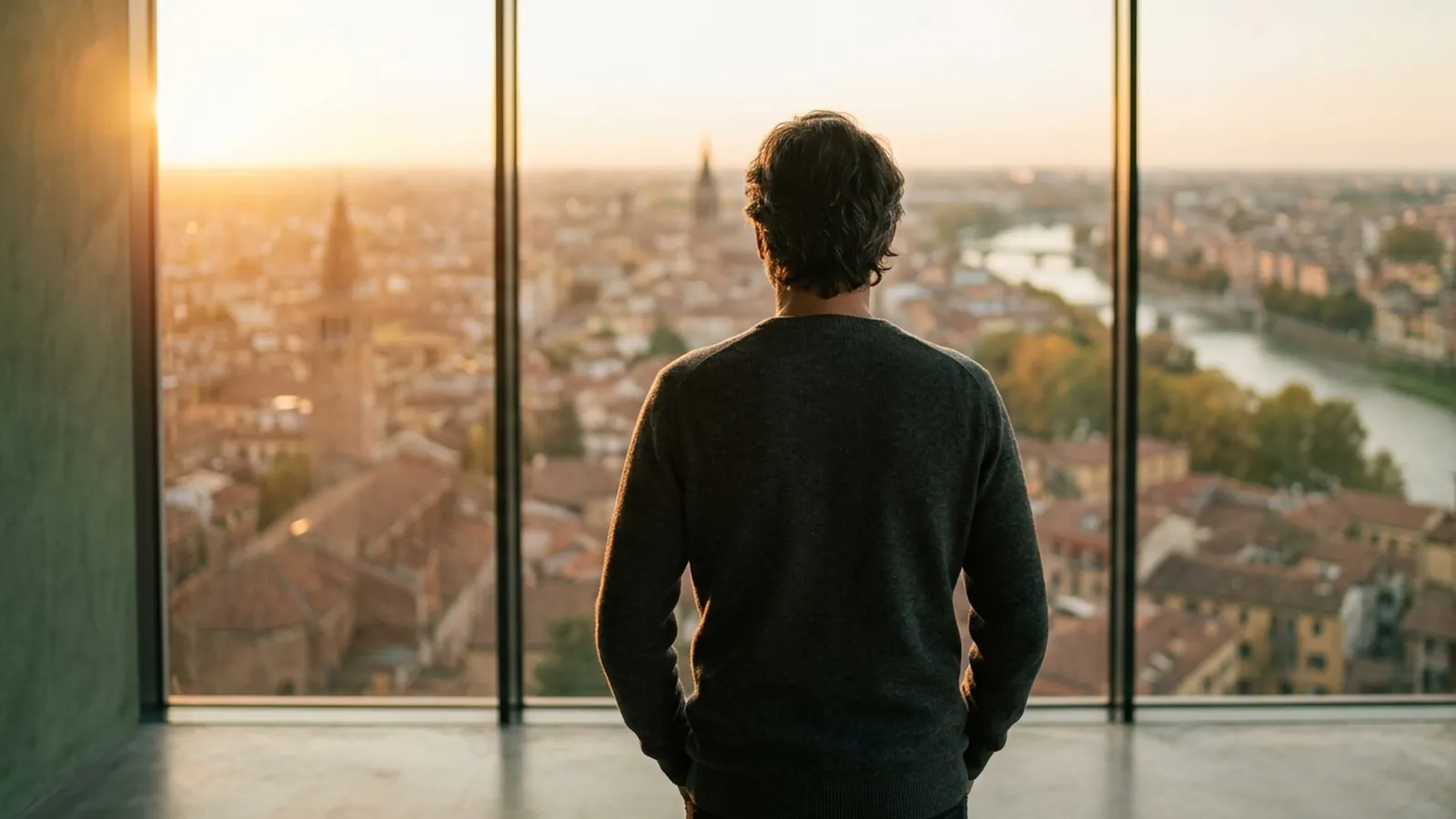 Un homme vu de dos, debout devant une grande baie vitrée, regarde une ville traversée par une rivière dans une lumière dorée
